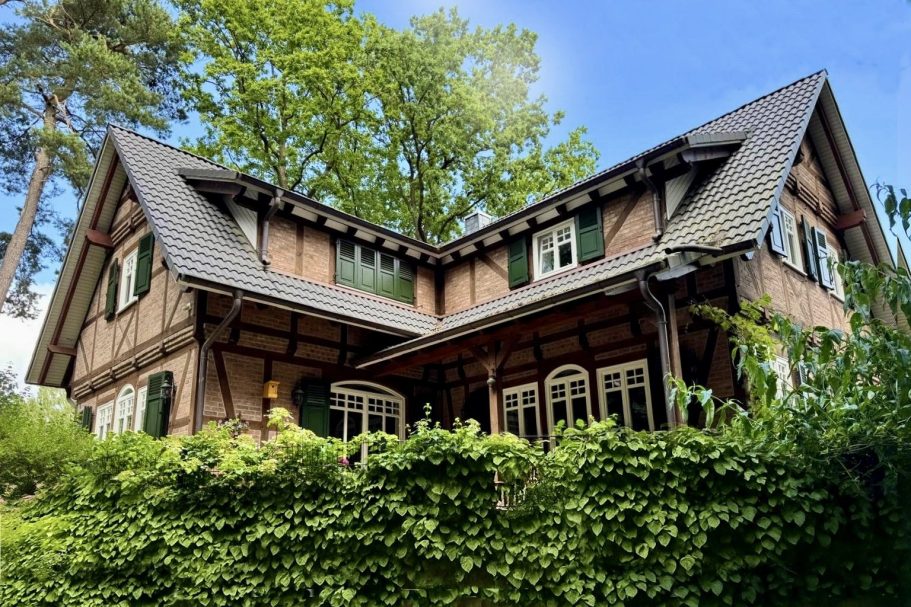 Historisches anmutendes Haus mit grünen Fensterläden, umgeben von Bäumen und Pflanzen.