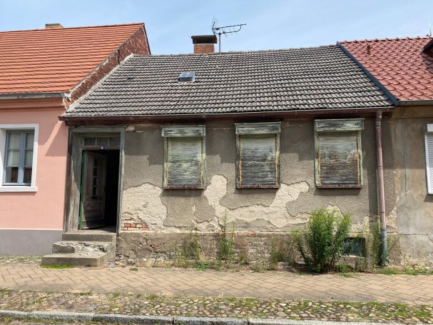 Verfallenes Haus mit mehreren Fenstern und abgeblätterter Fassade, umgeben von Gras.