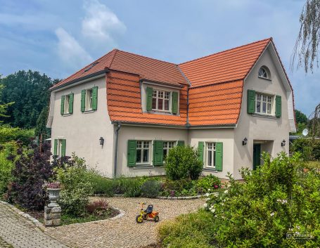 Zweifamilienhaus mit rotem Dach und grünen Fensterläden, umgeben von Gartenflächen.