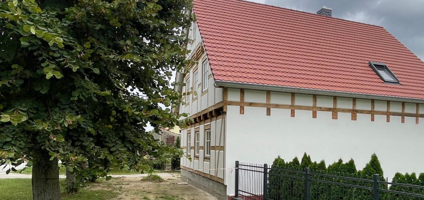 Fachwerkhaus mit hellem Putz mit rot gedecktem Dach und Blick auf einen Baum im Vordergrund.