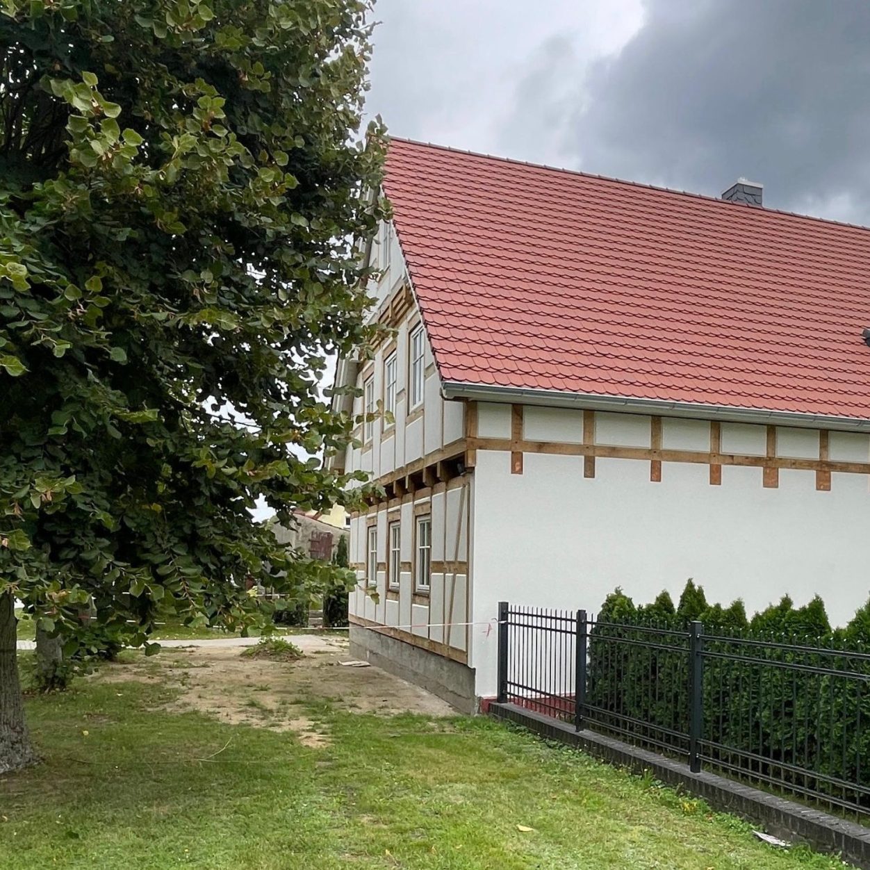 Ein zweigeschossiges Gebäude mit rotem Dach und Fachwerkelemente im Giebel, umgeben von Bäumen und einem grünen Garten.