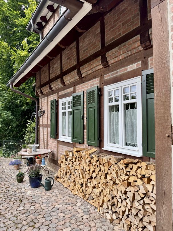 Holzstapel neben einem gemauerten Haus mit grünen Fensterläden und blühenden Töpfen.