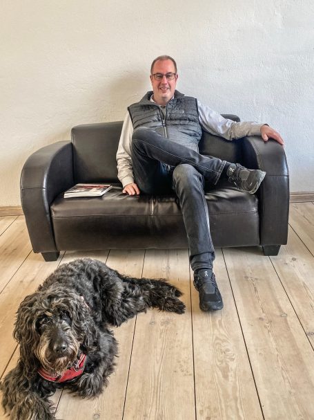 Mann sitzt auf einem Sofa, Hund liegt entspannt auf dem Boden neben ihm.