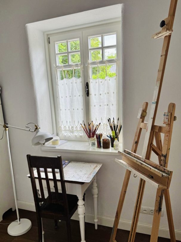 Helles Zimmer mit einem Tisch, Stuhl, Staffelei und Fenster, besetzt mit Künstlermaterialien.