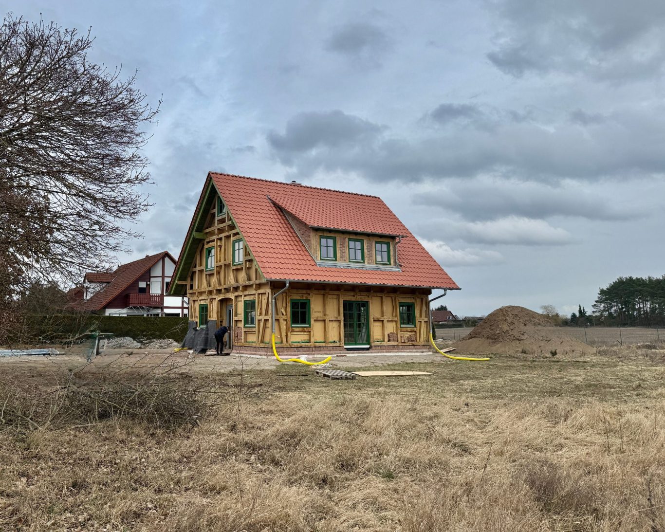 Ein freistehendes Fachwerkhaus im Rohbauzustand mit rotem Dach, umgeben von Wiese und Bäumen.