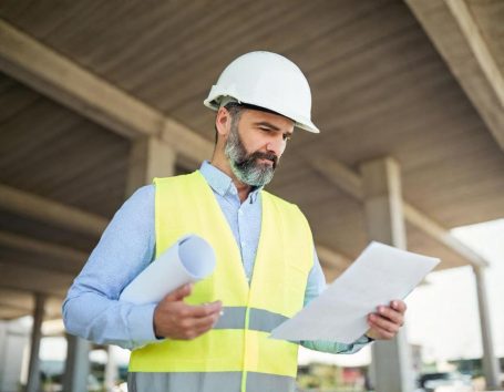 Mann mit sicherer Weste und Helm, der Baupläne auf einer Baustelle prüft.
