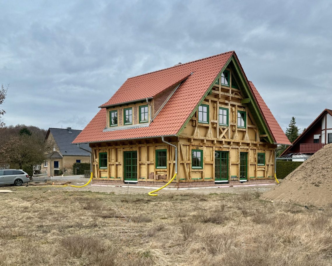 Neues Fachwerkhaus mit rotem Dach, grünem Rahmen und unbebautem Grundstück.
