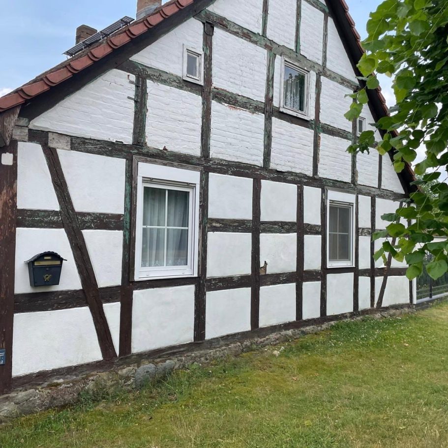 Fachwerkhaus mit weißer Fassade und braunen Balken, umgeben von Gras und Bäumen.