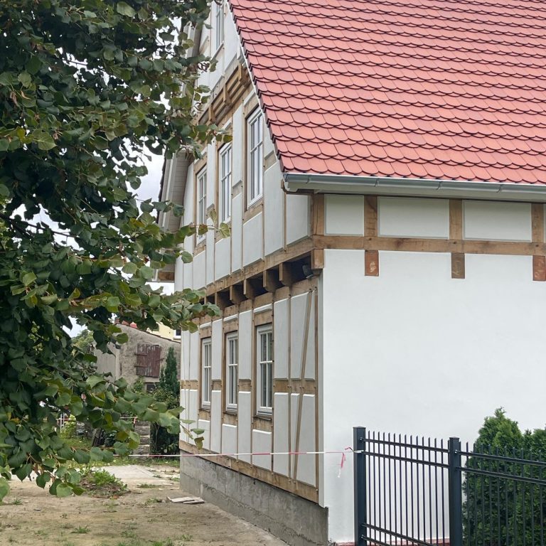 Fachwerkhaus mit rotem Satteldach und weißen Wänden, umgeben von Garten und Zaun.