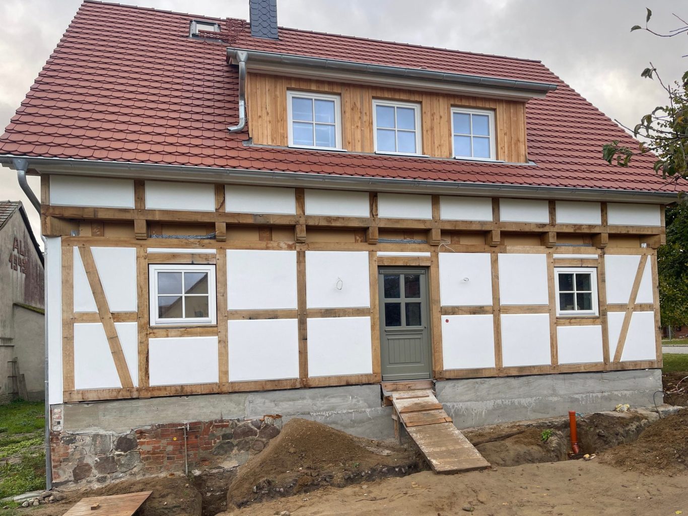 Fachwerkhaus mit hellem Putz mit rot gedecktem Dach und Blick auf einen Baum im Vordergrund.