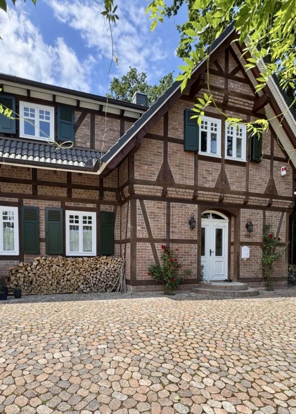 Fachwerkhaus mit grünen Fensterläden und gepflastertem Weg, umgeben von Bäumen.